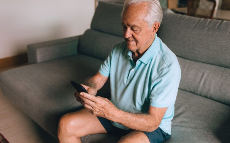 Man sitting on a grey sofa scrolling through recommended videos on his smartphone