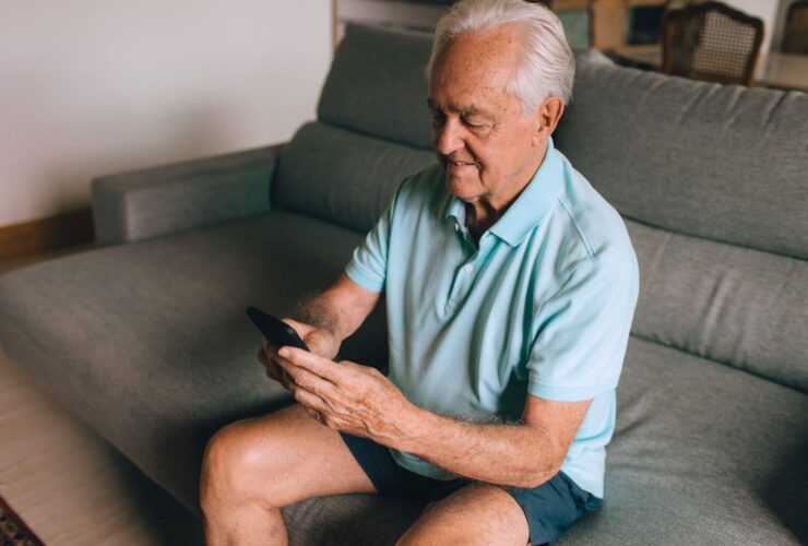 Man sitting on a grey sofa scrolling through recommended videos on his smartphone